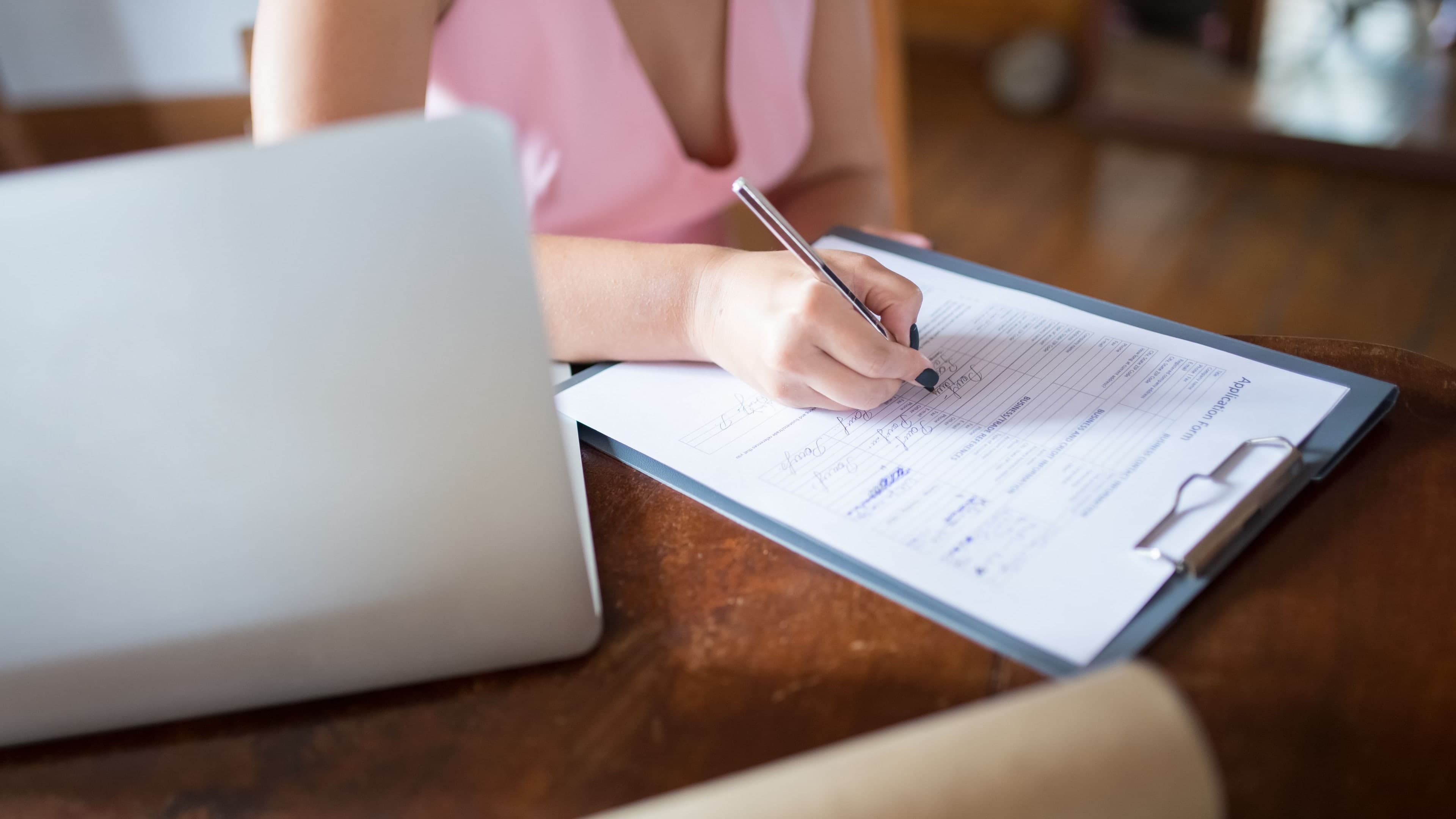 A woman fills in a document on a clipboard at a wooden desk, a laptop open beside her.