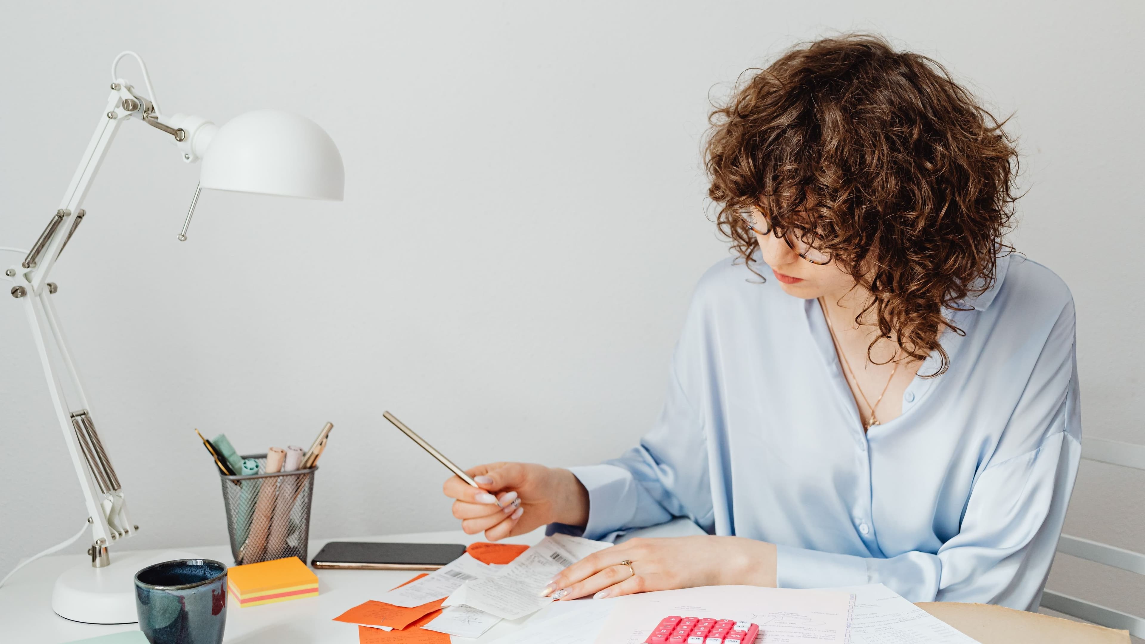 A woman at a wooden desk goes through receipts with a calculator and a cup of coffee beside her.