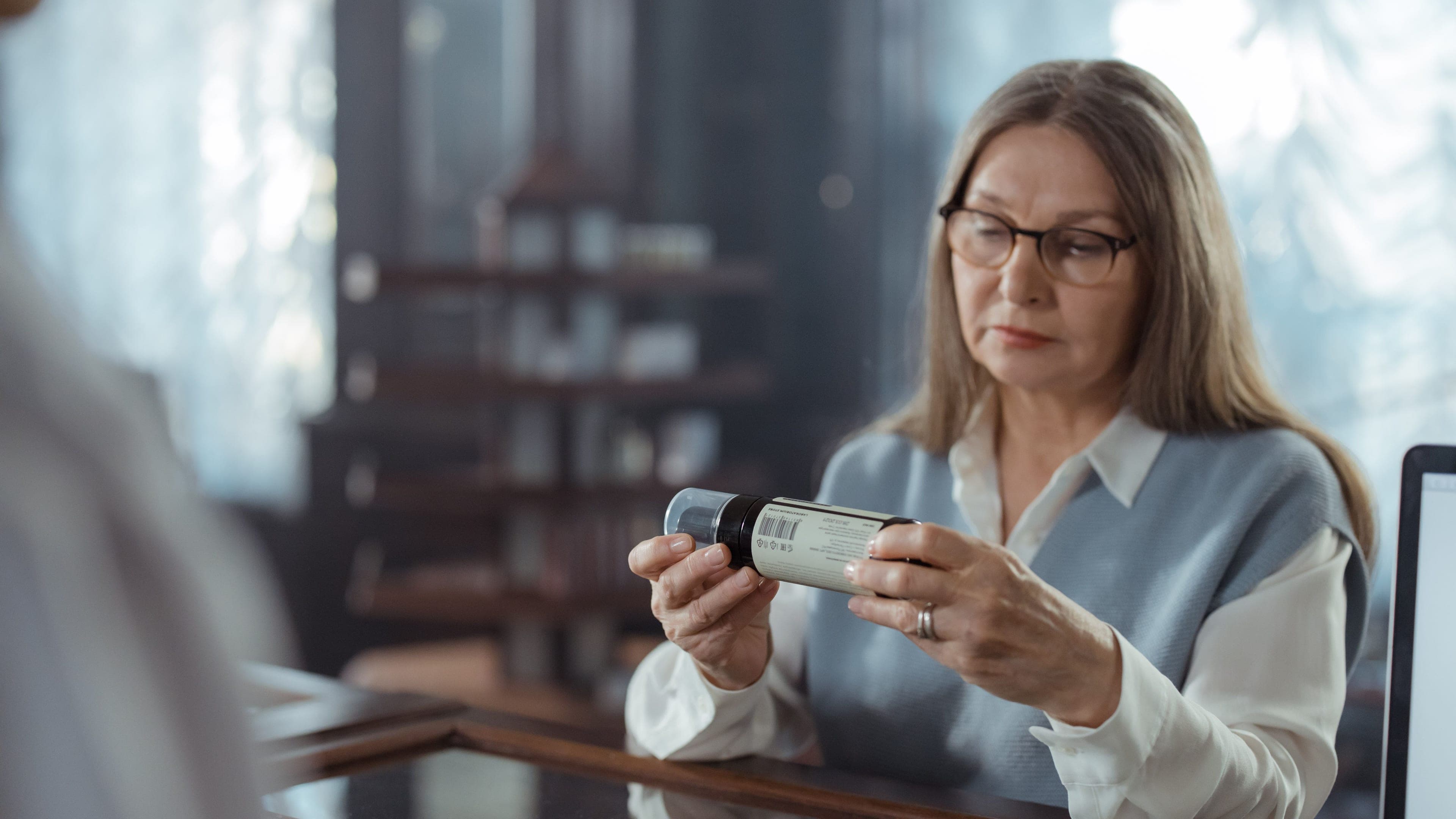 A practitioner with glasses reads the label on a supplement bottle at a wooden counter.