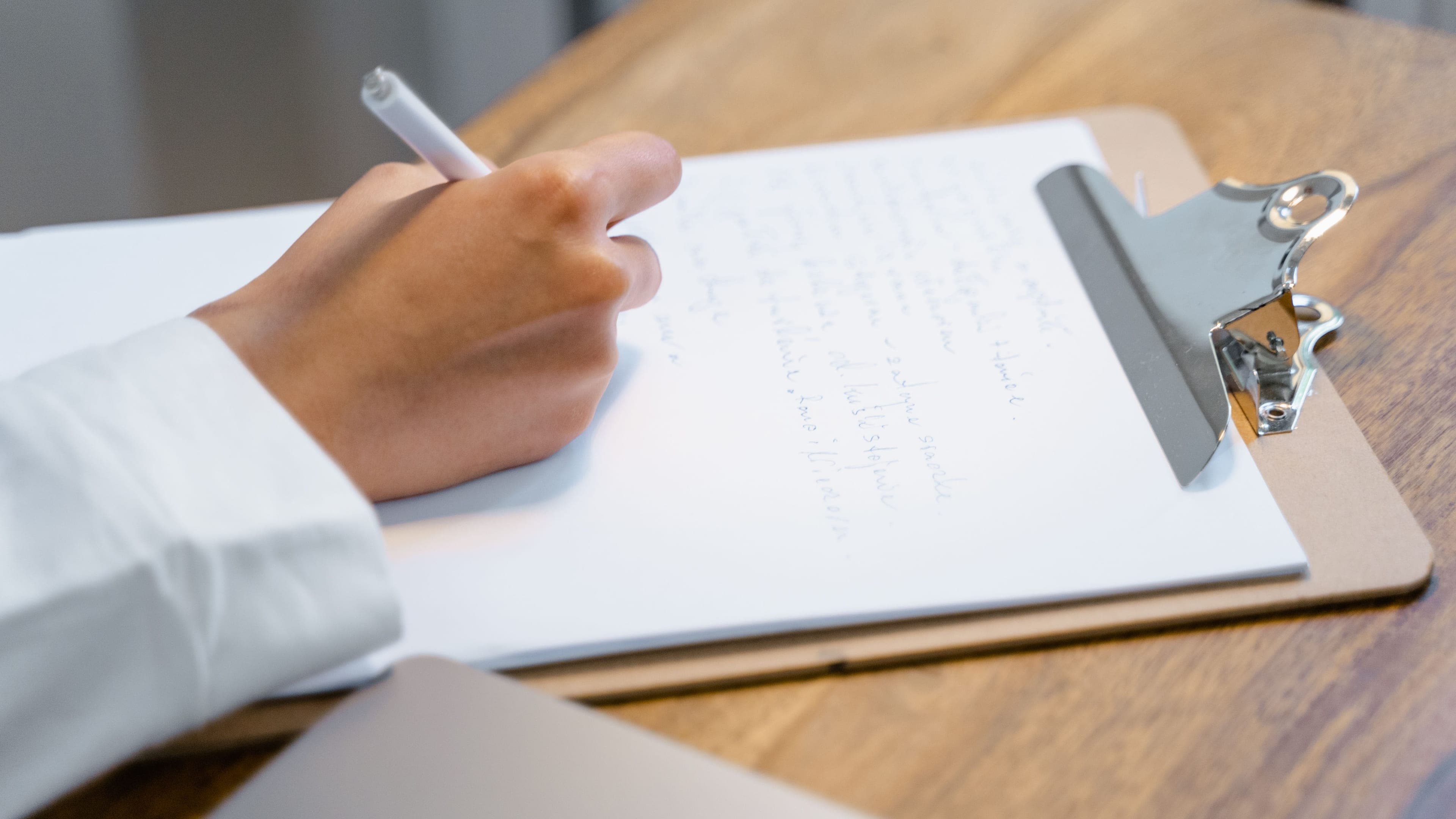 A hand writing with a pen on a paper form on a clipboard at a wooden desk.