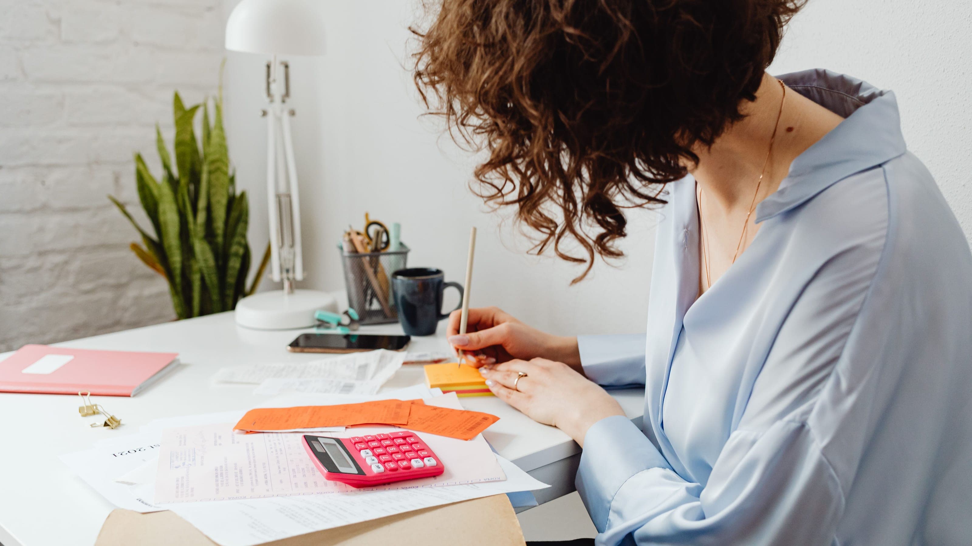 A woman at a white desk writes a note next to a pink calculator and a stack of receipts.