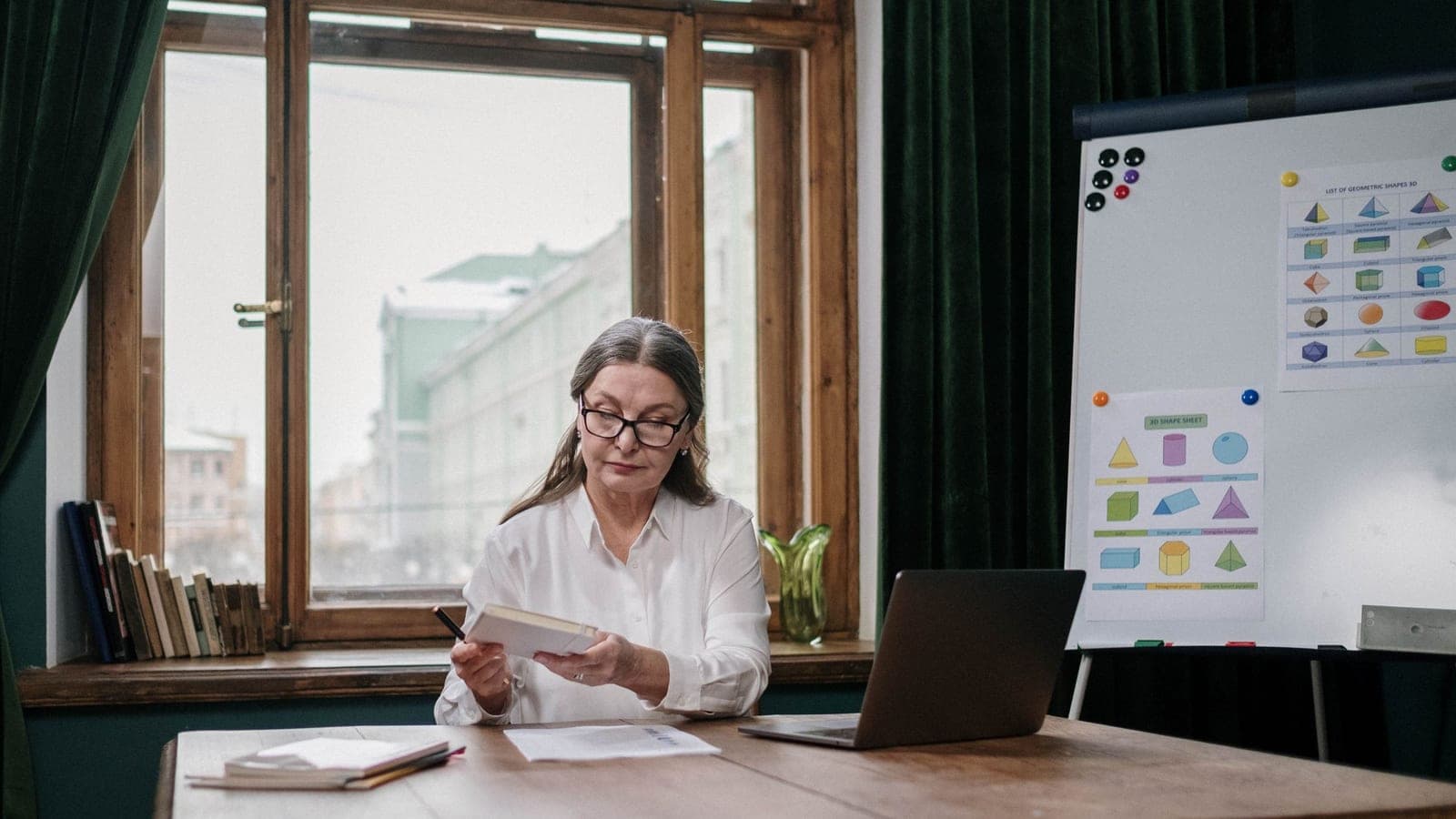 A practitioner with glasses reads through notes at a wooden desk, a laptop open beside her.