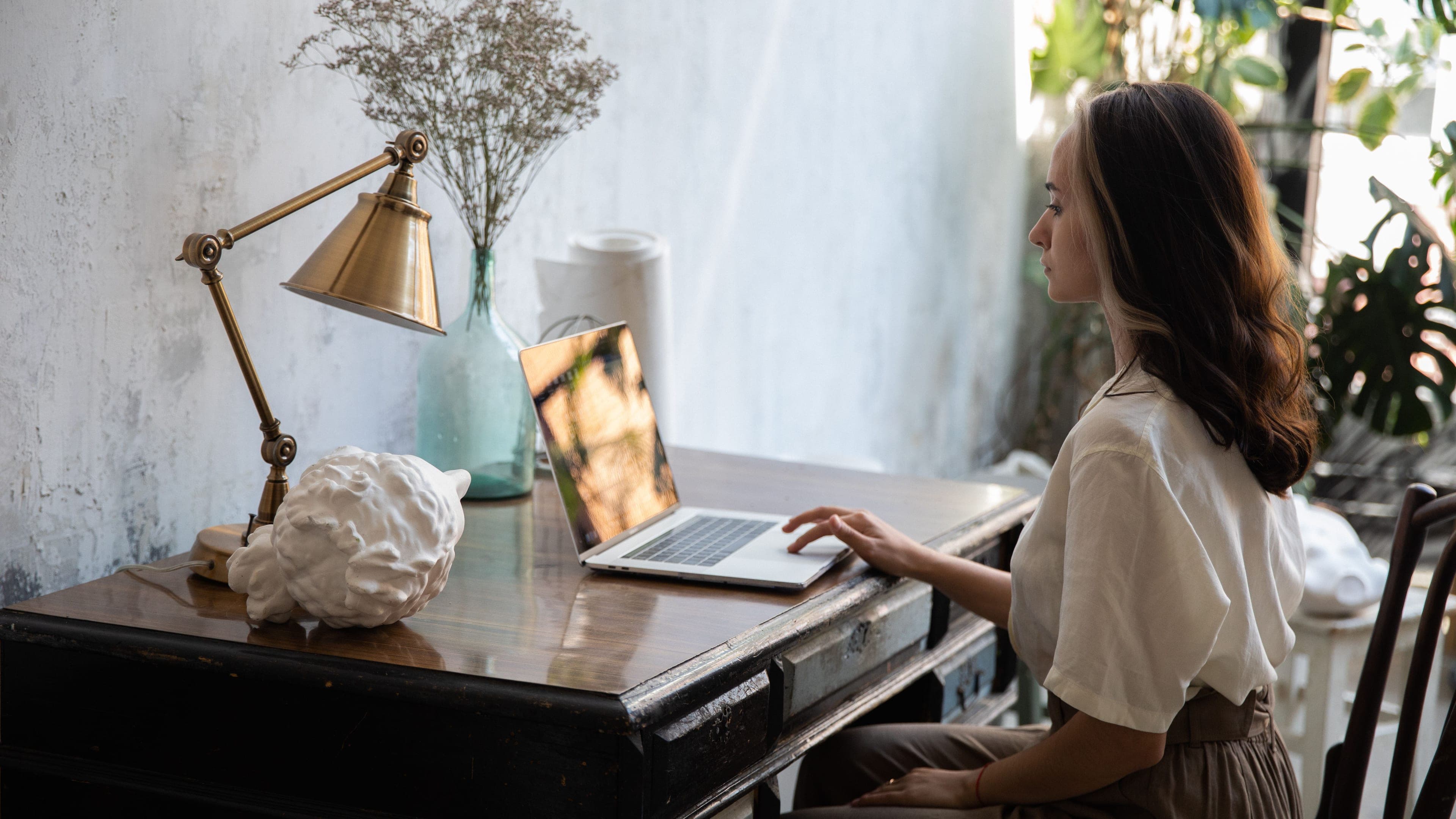 A woman works on a laptop at an old wooden desk with a brass lamp and dried flowers, plants in the light behind her.