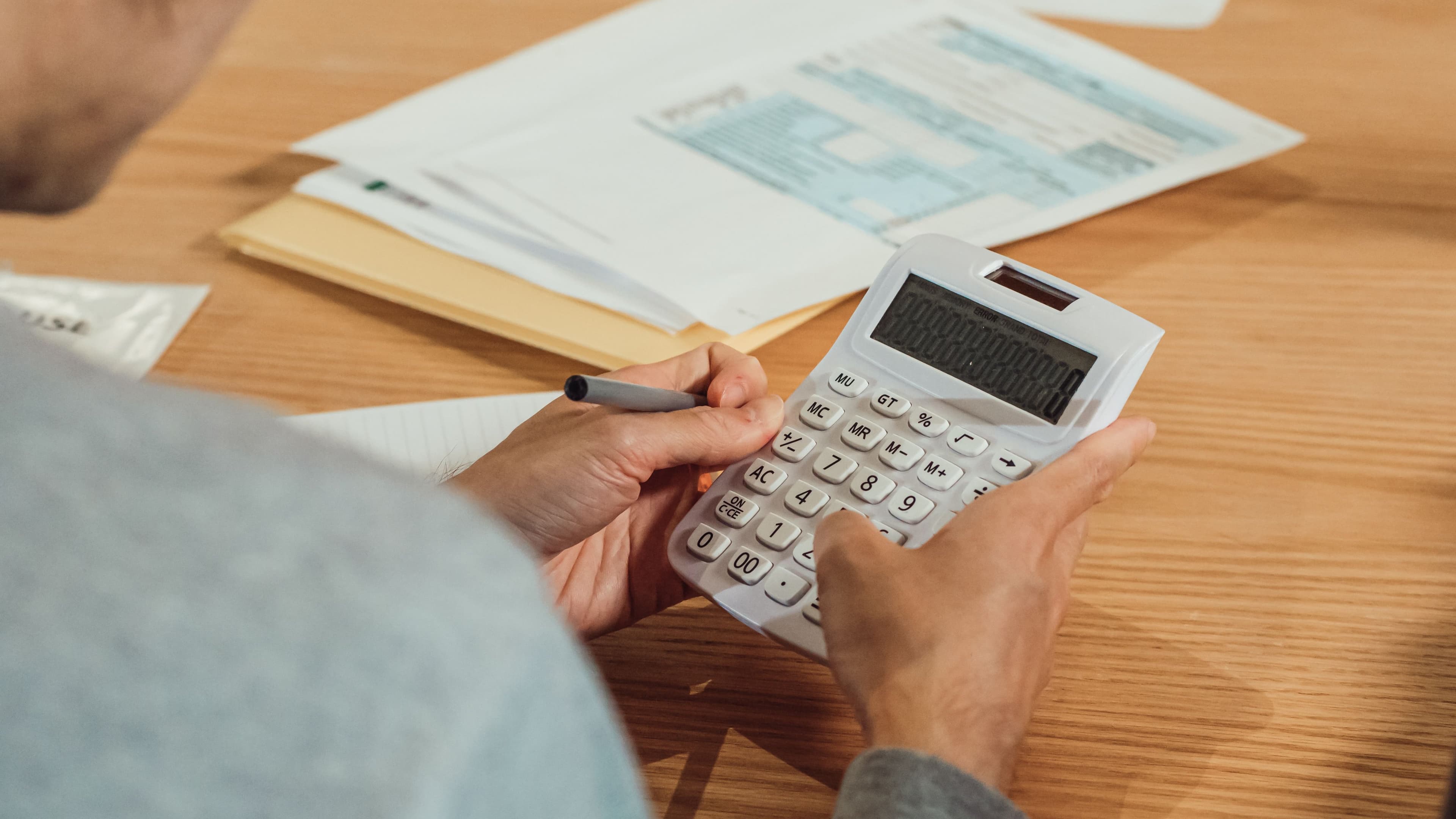A person works through papers with a calculator and a pen at a wooden desk.
